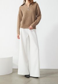 Femme portant un pull côtelé beige à col zippé, un pantalon blanc à jambes larges et des chaussures noires pointues, debout sur un sol en béton.