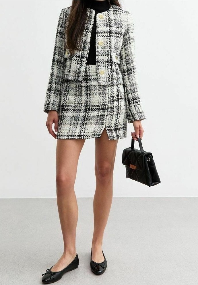 Femme portant une veste à carreaux noir et blanc et une jupe assortie, tenant un petit sac à main noir, portant des ballerines noires.