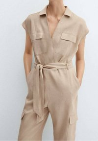 Mono de lino beige con mangas cortas, cuello, dos bolsillos en el pecho y cinturón atado en la cintura. Textura suave y corte relajado.