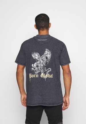 Maglietta in tessuto grigio con una grande grafica di un'aquila bianca e la scritta "Born Global" in giallo sul retro. Vestibilità comoda, maniche corte.