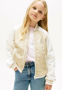 Chaqueta bomber beige con mangas blancas, puños y cuello acanalados. Parte delantera con cremallera, bolsillos laterales y detalle sutil del logo en el pecho. Llevada sobre una camisa rosa.