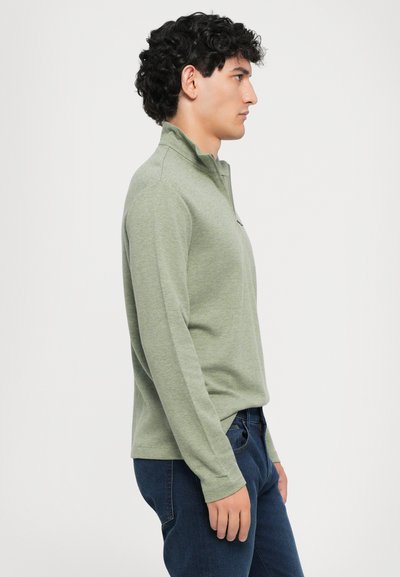 Zöld hosszú ujjú pulóver gallérral, texturált anyagból. Sötétkék farmernadrággal párosítva, oldalnézetből látható.