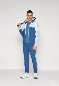 Blå og hvit treningsjakke med hette, kombinert med matchende blå bukser. Tilbehør inkluderer en tennisracket og hvite joggesko.