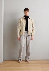 Beige lett jakke over en svart genser med høy hals, grå bukser og beige sko. Glatt stoff, avslappet passform, minimal hardware. Bakgrunn av tregulv.