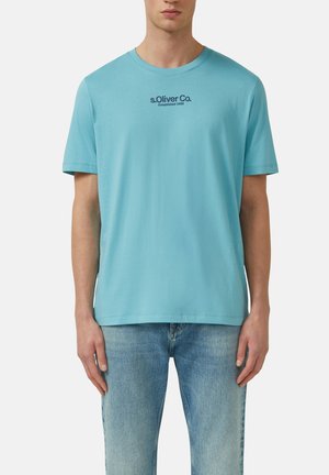 Mężczyzna ubrany w jasnoniebieską koszulkę s.Oliver Co. z krótkim rękawem i wyblakłe niebieskie dżinsy, stojący na jednolitym białym tle.