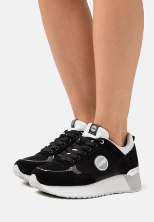 Baskets Colmar en noir et blanc portées aux pieds, avec détails en suède et en maille, logo rond sur le côté, semelle blanche épaisse avec soutien au talon.