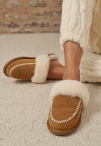 Le pantofole scamosciate color sabbia presentano un bordo in pelliccia sintetica color crema e un motivo di cuciture bianche decorative. Le suole sono piatte e marroni, con una texture morbida.