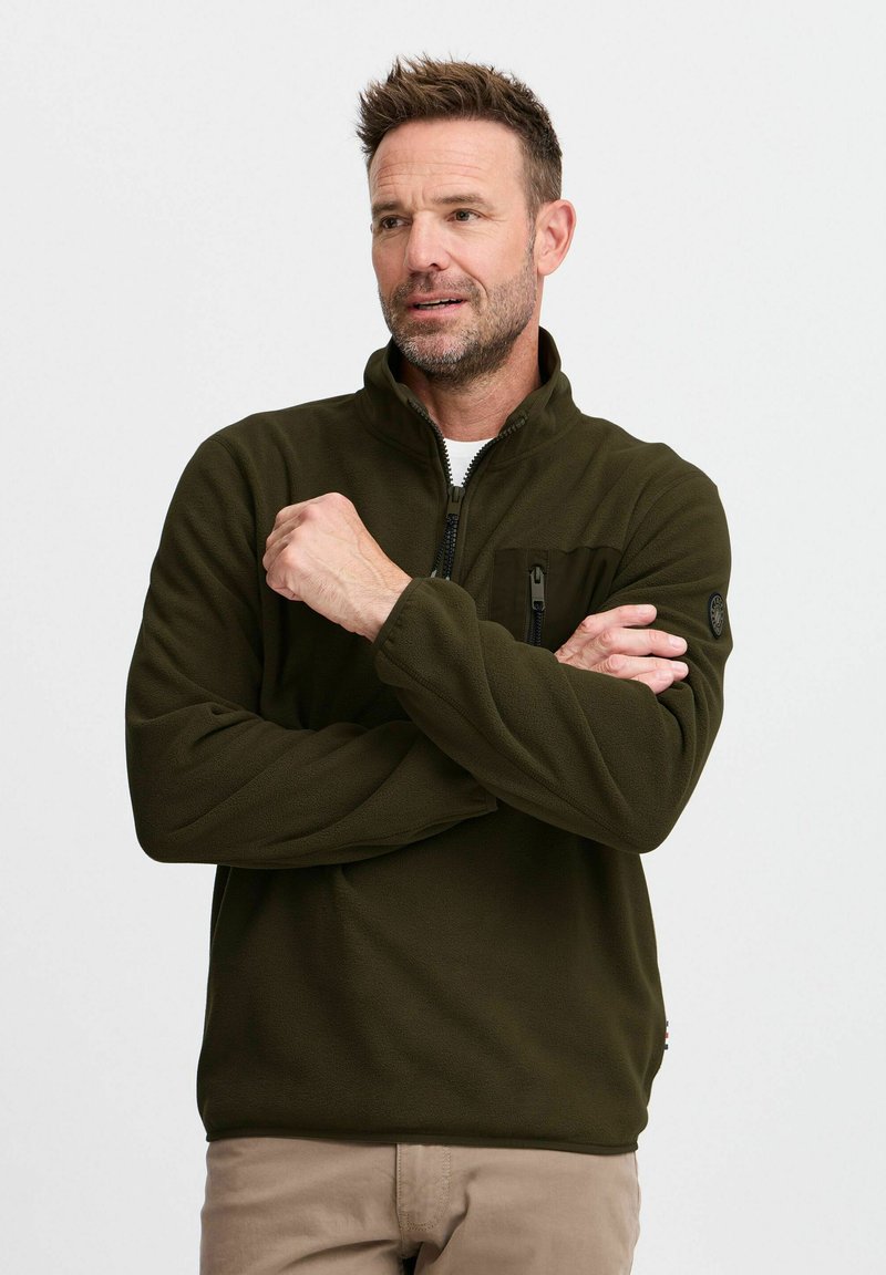 Pull en polaire vert olive avec un col zippé, une poche poitrine zippée et un badge sur l'épaule ; texture douce et coupe décontractée.
