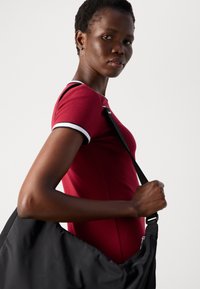 Woman wearing hoop earrings and a red short-sleeve top holding a large black shoulder bag against a plain light background.