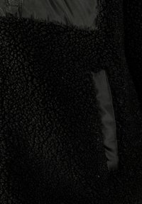 Veste en polaire noire avec une surface texturée et duveteuse, agrémentée d'accents en tissu lisse. Le design est chaud et pratique pour les temps froids.