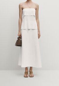 Top bianco a coste senza spalline sovrapposto a una lunga gonna bianca. Accessoriato con una piccola borsa marrone e sandali slipper beige.