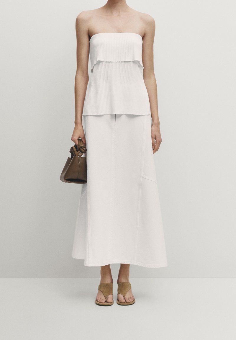 Top bianco a coste senza spalline sovrapposto a una lunga gonna bianca. Accessoriato con una piccola borsa marrone e sandali slipper beige.