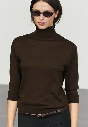 Donna che indossa occhiali da sole tartarugati, un maglione dolcevita marrone scuro con maniche a tre quarti e una cintura scura sopra pantaloni neri.