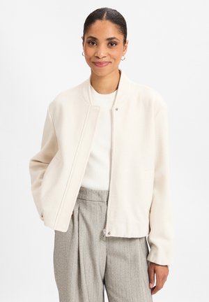 Jonge vrouw met hoepeloorbellen draagt een crème zip-up jas, een wit geribbelde top en grijze hoge taille broek met krijtstrepen, glimlacht zachtjes.