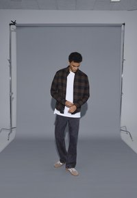 Karo srajca v črno-rjavi barvi čez belo majico, v kombinaciji s temnosivimi hlačami in sivimi supergami, stoji ob sivi ozadju.