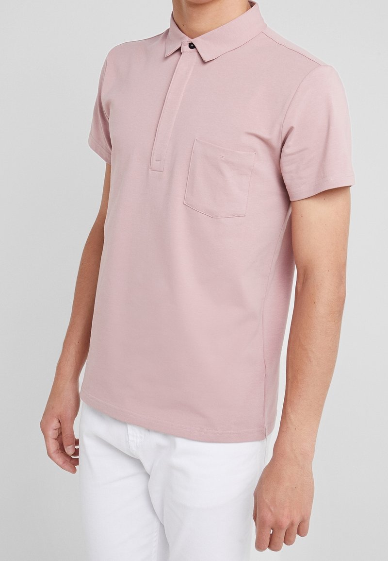 Homme portant un polo à manches courtes rose pâle avec une poche poitrine et un pantalon blanc, sur fond uni gris clair.