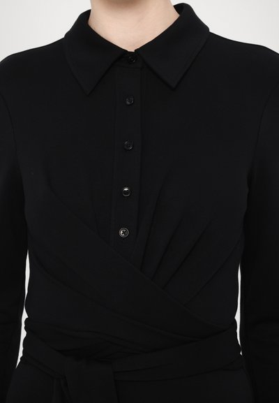 Robe noire avec un col, présentant une patte de boutonnage, un design croisé et un détail de taille nouée. Texture lisse sans motifs visibles.