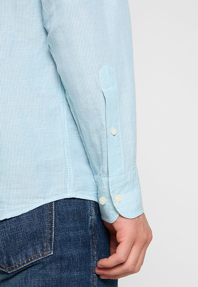 Himmelblau gestreiftes Hemd mit geknöpftem Manschette, aus weichem Stoff hergestellt. Sichtbare Nahtdetails und eine glatte Textur, kombiniert mit dunklen Jeans.