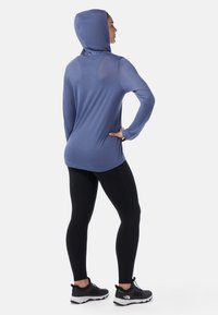 Long-sleeve, hooded top in light blue, made of soft fabric. Black leggings and athletic shoes complete the outfit, with a fitted silhouette.