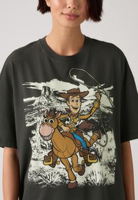 Camiseta oversized de color gris oscuro con una impresión de un vaquero de dibujos animados montando un caballo marrón, con un paisaje de fondo y un lazo en la mano.