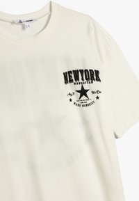 Cremefarvet t-shirt med korte ærmer og rund hals. Har et sort trykt "NEW YORK" grafik, stjerner og tekstdetaljer på forsiden.