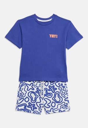 Camiseta azul de manga corta con texto "YAY!" en naranja combinada con pantalones cortos blancos con patrones abstractos en azul y cordón blanco.