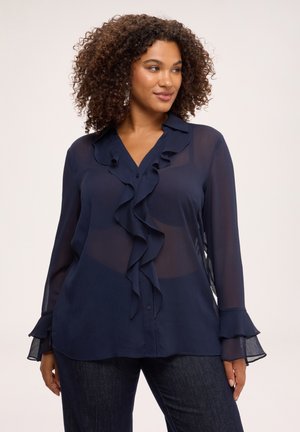 Frau mit lockigem Haar, die eine transparente marineblaue Bluse mit Rüschen vorne und an den Manschetten sowie dunkle Jeans trägt und vor einem einfarbigen Hintergrund steht.
