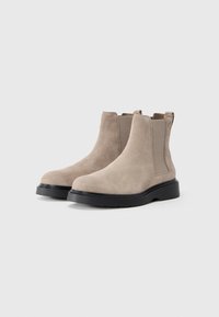 Bottines en suede beige avec des panneaux élastiques sur les côtés et une épaisse semelle en caoutchouc noir, présentant un bout arrondi et un design minimaliste.