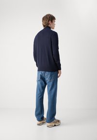 Navy pullover with a high collar, paired with light blue relaxed-fit jeans and beige sneakers featuring dark accents. Smooth texture.