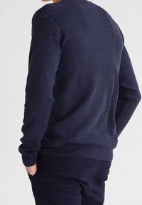 Pull en maille bleu marine avec un motif texturé, col rond et poignets côtelés. Porté avec un pantalon foncé, affichant un look décontracté.