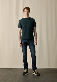 Dark green short-sleeve T-shirt with a round neckline, paired with fitted dark blue jeans and black, grey, and beige sneakers.