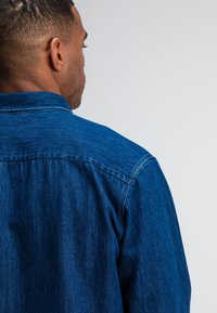 Donkerblauwe denim shirt met een standaardkraag, knoopsluiting en een relaxte pasvorm. De textuur van de stof lijkt zacht met een klassiek ontwerp.