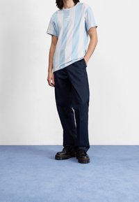 Světle modré a bílé pruhované bavlněné tričko s potiskem na přední straně, kombinované s tmavě modrými širokými kalhotami a černými platformovými botami, na modré podlaze.