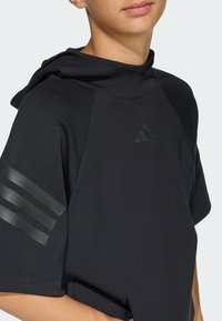 Ung person som bär en svart kortärmad hoodie med tre matta ränder på ärmen och en diskret logotyp på bröstet, närbild från hals till midja.