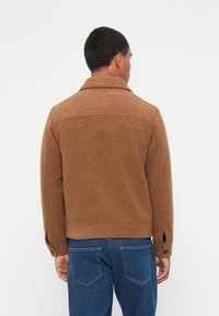 Giacca in misto lana marrone con colletto, polsini con bottoni e vestibilità dritta. Presenta una texture solida e un dettaglio allo yoke posteriore.