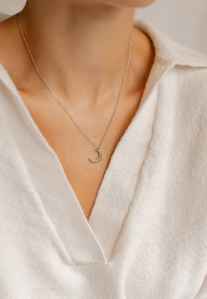Collier en chaîne en argent avec un petit pendentif en forme de croissant de lune d'une couleur verte douce, porté sur un vêtement à col blanc.