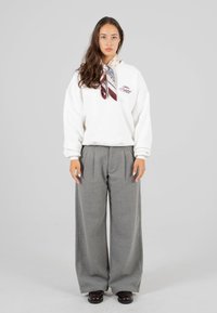 Una giovane donna sta in piedi rivolta in avanti indossando un felpa bianca, pantaloni grigi a gamba larga, scarpe scure e una sciarpa a fantasia attorno al collo, su uno sfondo neutro.