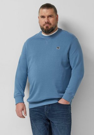 Mann mit Bart und zurückgebundenem Haar, der einen blauen Pullover und dunkle Jeans trägt und mit einer Hand in der Tasche vor einem einfarbigen Hintergrund steht.