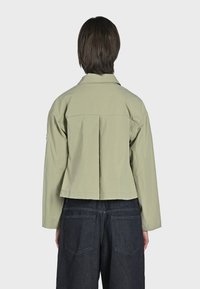 Ljust grön, kort jacka med en bakfåll, framkrage och långa ärmar, kombinerad med mörka jeans med bakfickor.