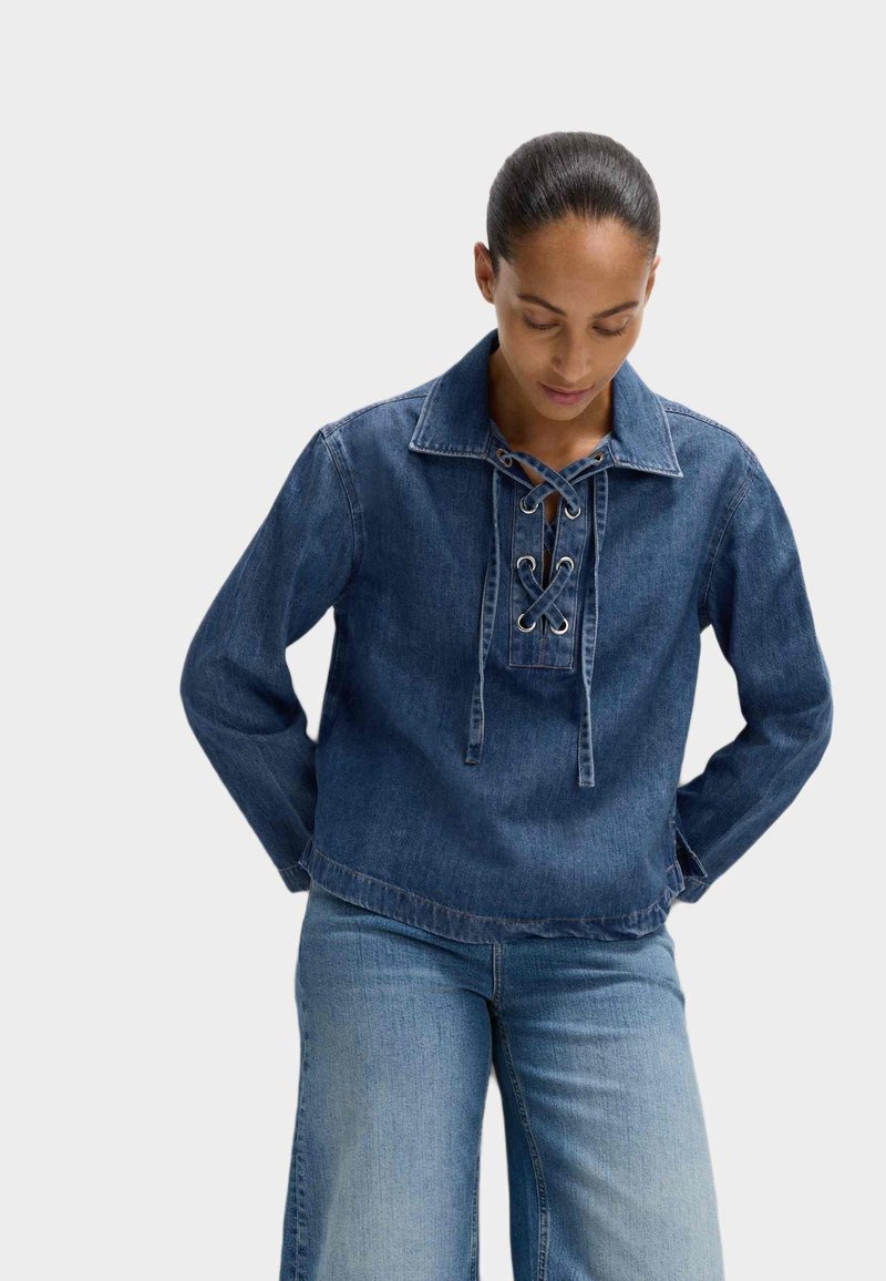 Pull en denim avec un décolleté lacé à l'avant, large col et manches longues. Couleur bleue avec une finition légèrement texturée. Longueur cropped et design décontracté.