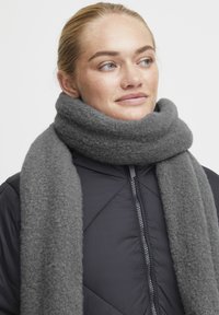 Grå ullskjerf pakket rundt halsen, med en myk tekstur. Skjerfet kontrasterer med en mørk, quiltet jakke.
