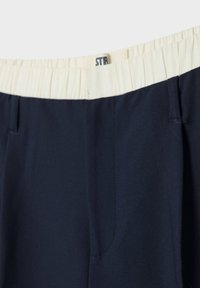 Pantalon bleu marine avec ceinture élastique blanche et étiquette partielle de la marque visible à l'intérieur de la ceinture.