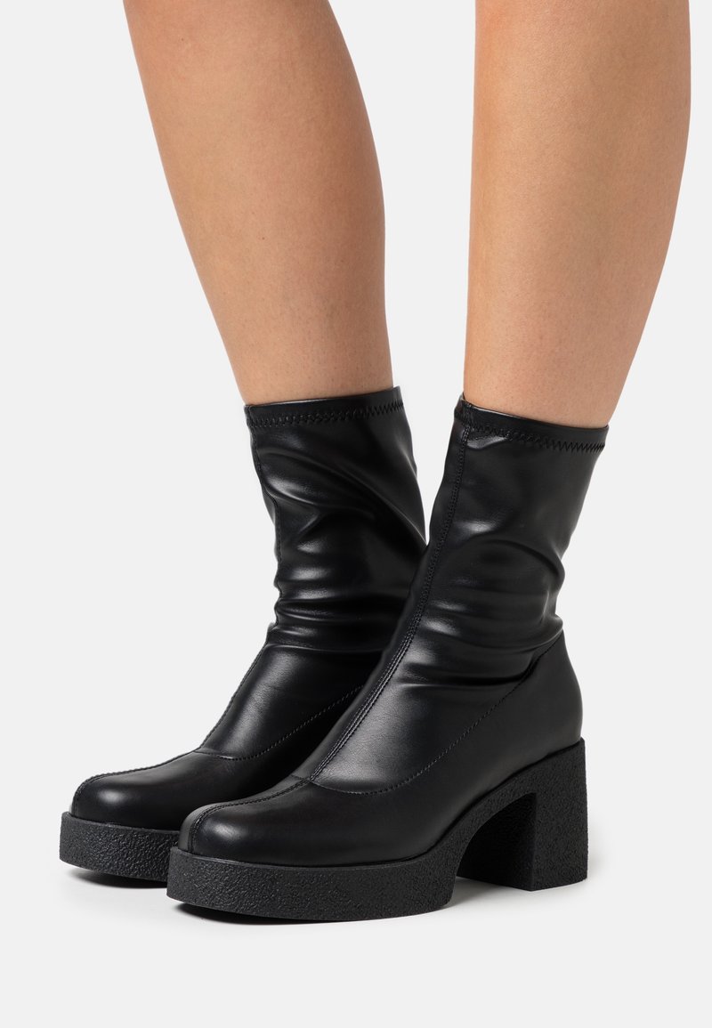 Bottes en cuir noir brillant mi-mollet avec talons blocs épais et texturés, portées sur des jambes nues sur un fond uni.