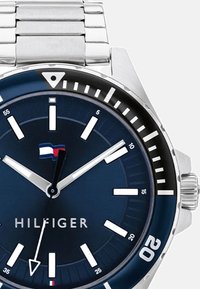 Reloj de acero inoxidable con una esfera azul marino, marcadores de hora blancos en negrita y un bisel negro. Las características incluyen una pulsera plateada y un logotipo a las 12 en punto.