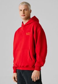 Joven con cabello rubio corto, vistiendo una sudadera con capucha roja de "Red Bull Racing" y pantalones oscuros, de pie frente a un fondo gris liso.