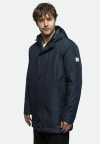 Marineblå vandtæt parka med hætte, frontlynlås, trykknapper og to sidelommer. Har et logo på venstre ærme.