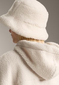 Osoba s svijetlo plavom kosom nosi kremasto obojeni teksturirani kapeć od flisa i odgovarajuću jaknu s kapuljačom, okrenuta lijevo protiv neutralne pozadine.