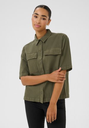 Žena s vlasmi zatiahnutými dozadu, oblečená v olivovo zelenú košeľu s krátkym rukávom a čierne nohavice, pózuje s jednou rukou prekríženou cez druhú.