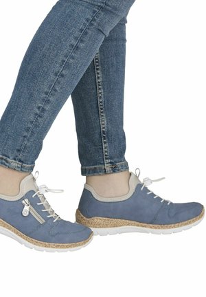 Nohy v modrých džínách, obuté v modrých sportovních teniskách s bílými podrážkami, béžovým lemováním, tkaničkami a bočním zipem, na bílém pozadí.