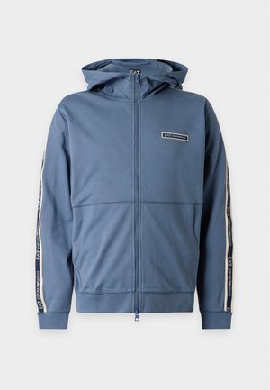 Homme portant un sweat à capuche bleu zippé avec des rayures sur les manches, un pantalon noir et des baskets blanches, debout devant un fond blanc uni.
